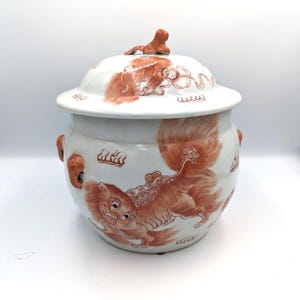 Puede incluir: Jarra de cerámica blanca con tapa, con ilustraciones rojas de criaturas míticas. La jarra tiene forma redonda, dos asas pequeñas y una tapa con una pequeña figura. La obra de arte es detallada y está sobre un fondo blanco.