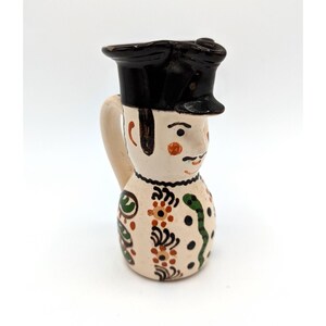 May include: A ceramic pitcher shaped like a person wearing a black hat with a white and brown face and a green and brown patterned body.