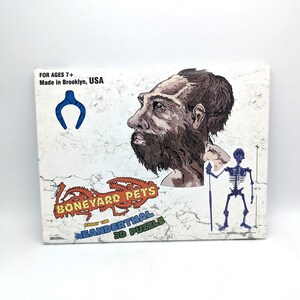 Peut inclure: Une boîte de puzzle avec le texte "BONEYARD PETS" et "HARRY THE NEANDERTHAL 3D PUZZLE". La boîte présente une illustration d'un homme de Néandertal et d'un squelette tenant une lance. La boîte est étiquetée "FOR AGES 7+" et "Made in Brooklyn, USA."