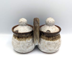 Puede incluir: Un juego de salero y pimentero de cerámica con asa. El juego presenta dos pequeños recipientes redondos con tapas. Los recipientes son blancos con detalles marrones y un asa marrón. Las tapas tienen pequeños agujeros.