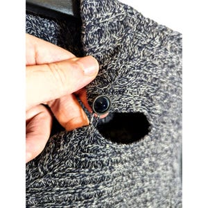 K&ouml;nnte beinhalten: Nahaufnahme eines grau-wei&szlig; gestrickten Pullovers mit dunklem Knopfverschluss. Der Pullover hat ein strukturiertes Aussehen und ein Knopflochdetail. Der Knopf hat eine dunkle, gl&auml;nzende Farbe.