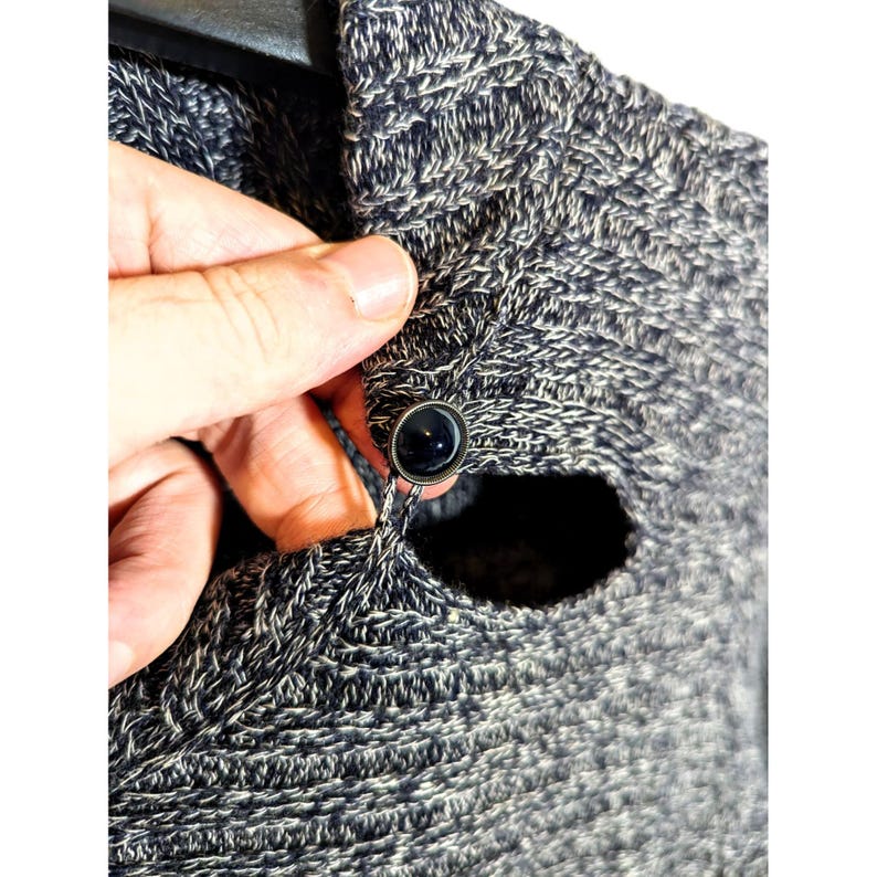 K&ouml;nnte beinhalten: Nahaufnahme eines grau-wei&szlig; gestrickten Pullovers mit dunklem Knopfverschluss. Der Pullover hat ein strukturiertes Aussehen und ein Knopflochdetail. Der Knopf hat eine dunkle, gl&auml;nzende Farbe.
