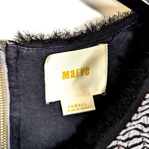 May include: Close-up of a black dress with a gold zipper and a label that reads "Maeve" in gold lettering. The label also indicates the size is XSMALL and "Made in China". The dress has a textured, fringed neckline.