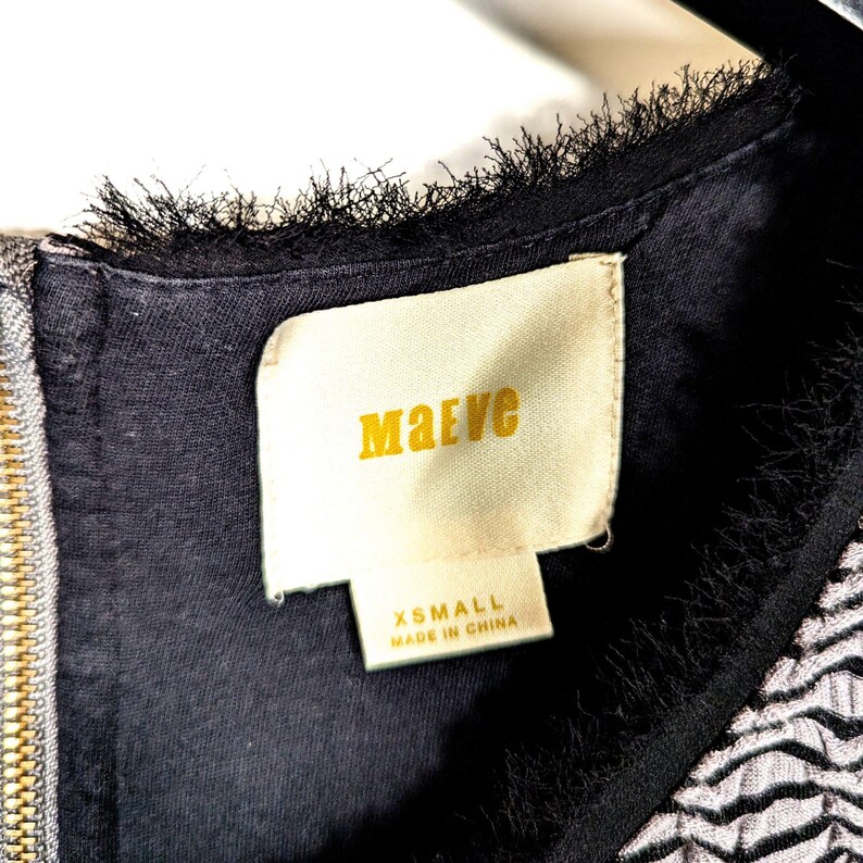 May include: Close-up of a black dress with a gold zipper and a label that reads "Maeve" in gold lettering. The label also indicates the size is XSMALL and "Made in China". The dress has a textured, fringed neckline.