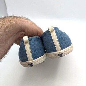 May include: A pair of blue denim slip-on shoes with cream accents and a Mickey Mouse silhouette on the heel. The shoes are held by a hand against a white background. The shoes are likely for children.