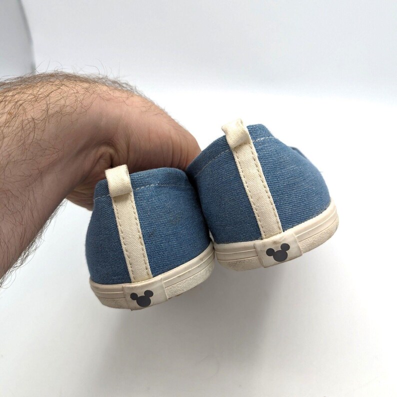 May include: A pair of blue denim slip-on shoes with cream accents and a Mickey Mouse silhouette on the heel. The shoes are held by a hand against a white background. The shoes are likely for children.