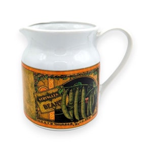 Puede incluir: Jarra de cerámica blanca con asa curva y boquilla. La jarra presenta una etiqueta de estilo vintage con las palabras "Stringless Beans" y una ilustración de judías verdes. La etiqueta tiene un borde naranja y dorado.