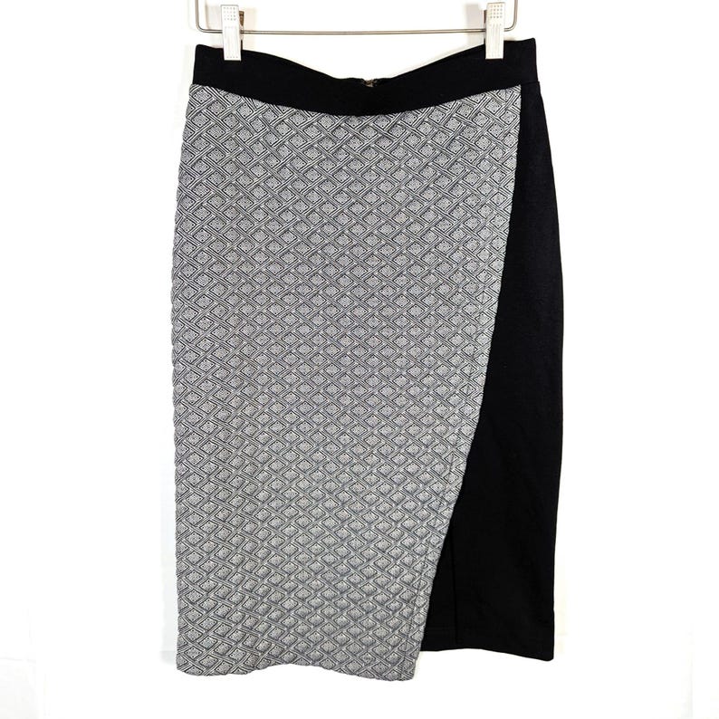May include: A black and white patterned pencil skirt with a black waistband and a side slit. The skirt is hanging on a white hanger against a white background. The skirt's pattern is a repeating geometric design.