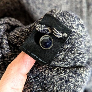 K&ouml;nnte beinhalten: Nahaufnahme eines marineblauen Knopfes mit silbernem Rand, befestigt an einem dunklen Stoffetikett. Das Etikett ist auf einem strukturierten, blau-wei&szlig; gestrickten Stoff befestigt. Der Text "MAIN FROID DE LAVAGE" ist sichtbar.