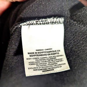 May include: Close-up of a tag with text. The tag reads "RN#56323-CA#05553 MADE IN EGYPT/FABRIQUE EN EGYPTE/HECHO EN EGIPTO". The tag also lists the materials used, including 100% polyester. The tag is white with black text.