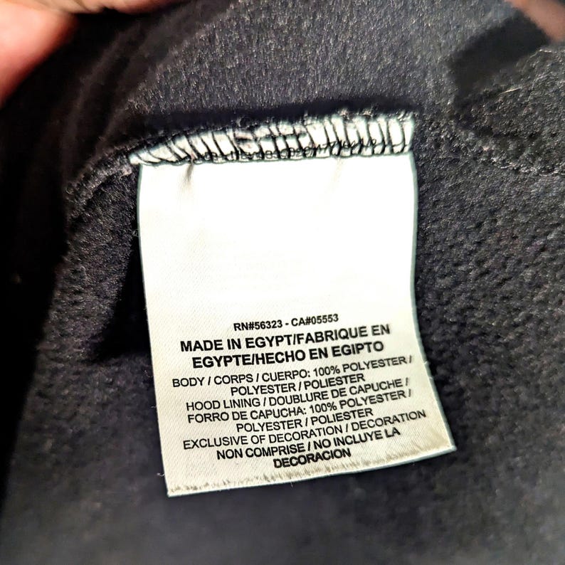 May include: Close-up of a tag with text. The tag reads "RN#56323-CA#05553 MADE IN EGYPT/FABRIQUE EN EGYPTE/HECHO EN EGIPTO". The tag also lists the materials used, including 100% polyester. The tag is white with black text.