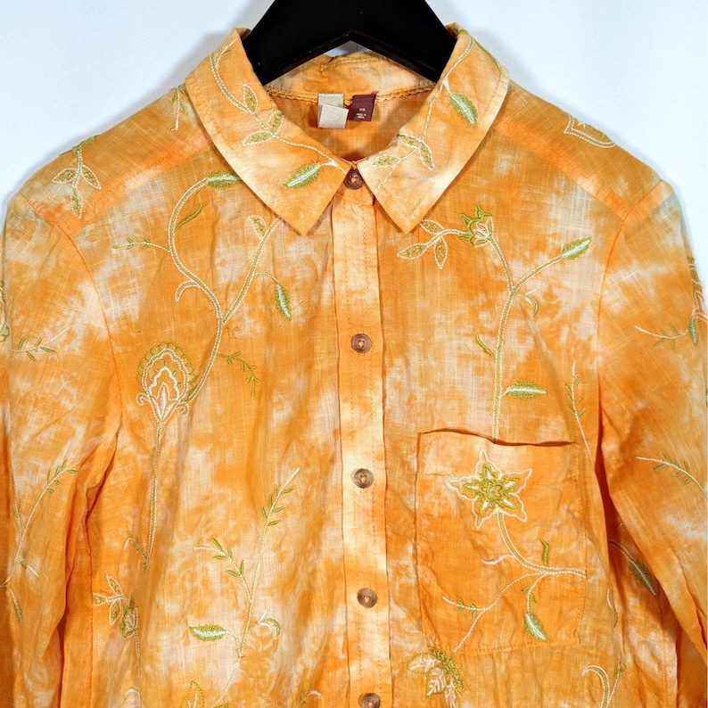 Puede incluir: Una camisa naranja con botones, con efecto tie-dye y patrones florales bordados. La camisa tiene cuello, bolsillo en el pecho y botones de madera. El bordado es en tonos verdes y blancos.