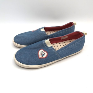 May include: Pair of blue denim slip-on shoes with a white sole. Each shoe features a heart-shaped patch with Mickey and Minnie Mouse. The interior lining is red with a small black and white pattern.