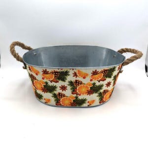 May include: Oval metal container with a decorative floral pattern featuring orange slices, pine needles, and cinnamon sticks. The container has rope handles and a silver interior, perfect for seasonal decor.