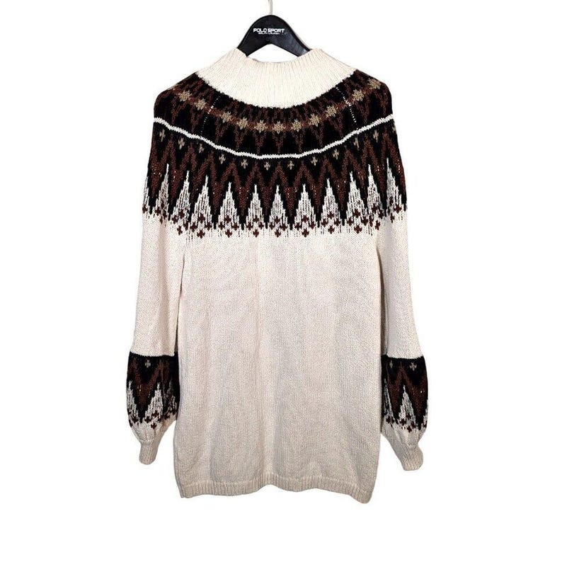 Puede incluir: Su&eacute;ter color crema con cuello alto y mangas largas. La parte superior del pecho y las mangas presentan un patr&oacute;n geom&eacute;trico en marr&oacute;n y blanco. El su&eacute;ter est&aacute; colgado de una percha negra.
