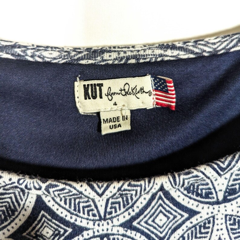 Puede incluir: Primer plano de una blusa azul marino con un estampado geom&eacute;trico blanco. La etiqueta dice "KUT from the Kloth" con una peque&ntilde;a bandera americana y "MADE IN USA". La blusa es talla 4.