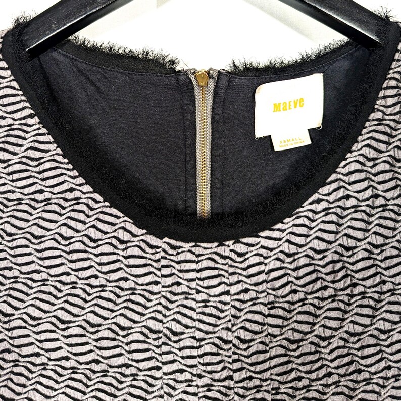 May include: Close-up of a black and white patterned dress. The dress has a black neckline with fringe detailing and a gold zipper. The tag reads "Maeve" and "XS/SMALL".