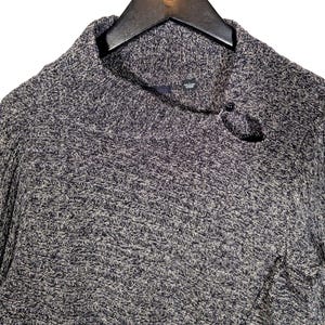 K&ouml;nnte beinhalten: Ein grau-blauer Strickpullover mit einem Rollkragen und einem Knopfverschluss. Der Pullover besteht aus einem strukturierten Strickstoff und h&auml;ngt an einem schwarzen Kleiderb&uuml;gel.