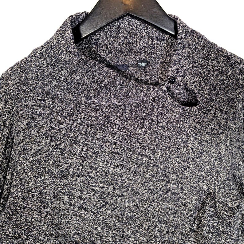 K&ouml;nnte beinhalten: Ein grau-blauer Strickpullover mit einem Rollkragen und einem Knopfverschluss. Der Pullover besteht aus einem strukturierten Strickstoff und h&auml;ngt an einem schwarzen Kleiderb&uuml;gel.