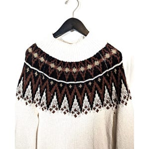 Puede incluir: Su&eacute;ter color crema con cuello alto y canes&uacute; estampado. El canes&uacute; presenta un dise&ntilde;o geom&eacute;trico en marr&oacute;n, negro y blanco. El su&eacute;ter est&aacute; colgado de una percha negra sobre un fondo blanco.