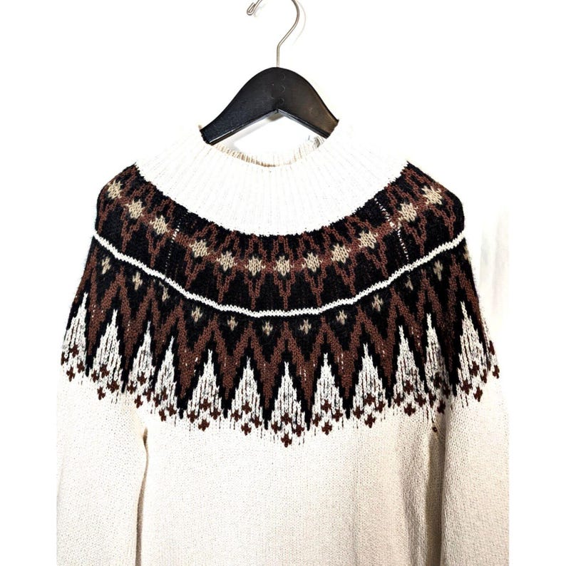 Puede incluir: Su&eacute;ter color crema con cuello alto y canes&uacute; estampado. El canes&uacute; presenta un dise&ntilde;o geom&eacute;trico en marr&oacute;n, negro y blanco. El su&eacute;ter est&aacute; colgado de una percha negra sobre un fondo blanco.