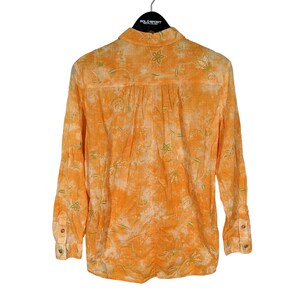 Puede incluir: Camisa naranja de manga larga con bordado floral. La camisa presenta un dise&ntilde;o abotonado y un cuello cl&aacute;sico. El bordado floral es en verde y blanco, contrastando con la tela naranja. La camisa tiene un aspecto texturizado.