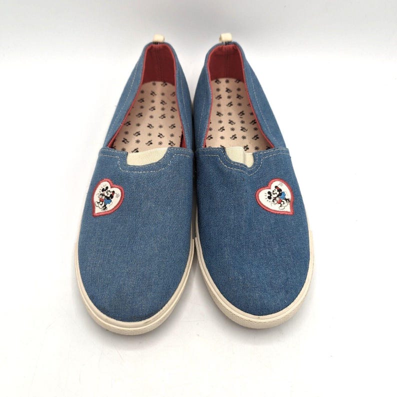 May include: A pair of blue denim slip-on shoes with white soles. Each shoe has a heart-shaped patch with a Mickey Mouse design. The interior lining is patterned with small, repeating shapes. The shoes have a rounded toe and a low profile.