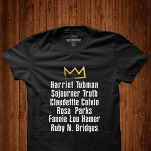 Könnte beinhalten: Schwarzes T-Shirt mit einer goldenen Krone und einer Liste von Namen: Harriet Tubman, Sojourner Truth, Claudette Colvin, Rosa Parks, Fannie Lou Hamer, Ruby N. Bridges.