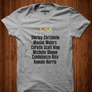 May include: Grey t-shirt featuring a gold crown and a list of names: Shirley Chrisholm, Maxine Waters, Coretta Scott King, Michelle Obama, Condoleezza Rice, Kamala Harris.