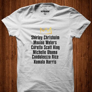 May include: White t-shirt featuring a gold crown and a list of names: Shirley Chrisholm, Maxine Waters, Coretta Scott King, Michelle Obama, Condoleezza Rice, Kamala Harris.