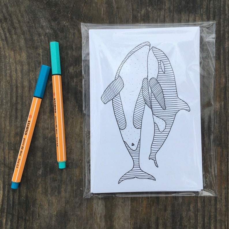 May include: A set of greeting cards featuring a black and white illustration of two orca whales, packaged in a clear plastic sleeve. Two orange and blue Stabilo pens are next to the cards.