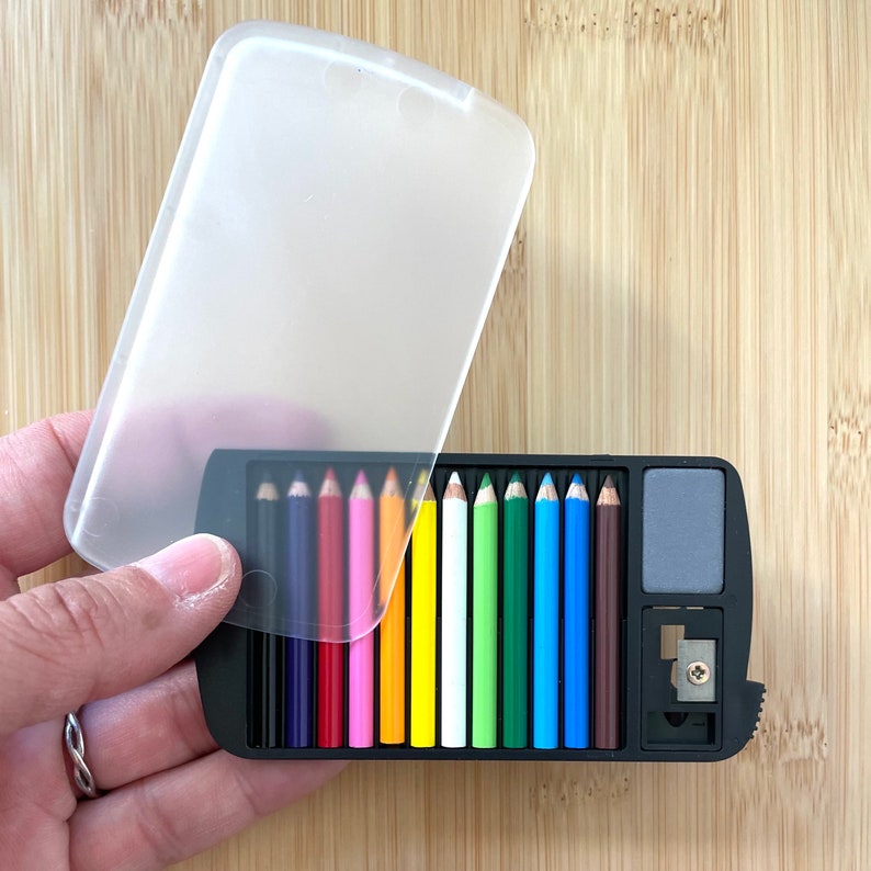 May include: A black pencil case with a clear lid, holding a set of twelve colored pencils. The pencils are arranged in a row, displaying a spectrum of colors. The case also includes a small eraser and a pencil sharpener.