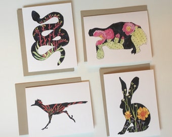 May include: Four greeting cards with animal illustrations. The cards feature a snake, a tortoise, a roadrunner, and a rabbit. Each animal is a black silhouette filled with colorful floral patterns. The cards have a white background and are paired with gray envelopes.