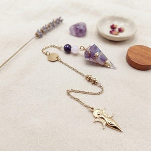 Può includere: Un pendolo in metallo dorato con una punta in cristallo sfaccettato viola e bianco. La catena include una piccola perlina viola scuro, una perlina rosa chiaro e un ciondolo dorato a forma di simbolo di luna crescente all'estremità.