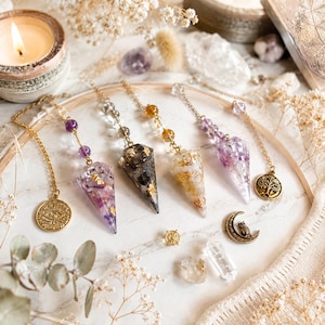 Peut inclure: Quatre pendules en résine pointus avec des flocons d'or, chacun attaché à une chaîne dorée. L'un comporte des perles d'améthyste violettes, un autre des perles transparentes, un des perles jaunes et un des perles violettes. Breloques : œil, arbre, hibou.