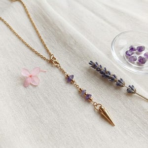 Könnte beinhalten: Eine goldfarbene Kettenhalskette mit vertikalem Anhänger-Design, besetzt mit kleinen violetten Amethyst-Splitterperlen und einem spitzen goldfarbenen Metallanhänger am Ende. Die Kette liegt neben einer einzelnen rosa Blüte und Lavendel.