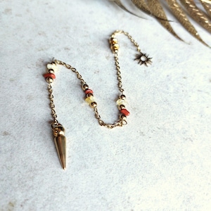 Può includere: Collana color oro con pendente e catena. La collana presenta un pendente a punta, un ciondolo a forma di sole ed è adornata con piccole perline rosse, gialle e bianche. Il gioiello è esposto su una superficie chiara.