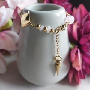 Könnte beinhalten: Ein goldfarbenes Armband mit einem geometrischen quadratischen Anhänger und einem hängenden kegelförmigen Anhänger. Das Armband hat kleine goldene Perlen und hellrosa Steinakzente. Der Schmuck wird auf einer weißen Keramikvase mit rosa Blumen im Hintergrund präsentiert.