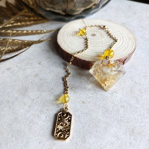 Könnte beinhalten: Ein goldfarbenes Pendel mit einem rautenförmigen Kristallanhänger, der mit Goldflocken gefüllt ist. Das Pendel hat eine Kette mit gelben Perlen und einen rechteckigen Anhänger mit einem Auge und einem Himmelsdesign. Das Pendel liegt auf einer Holzscheibe.