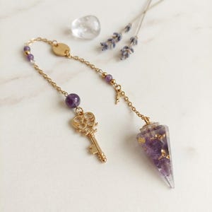 Könnte beinhalten: Ein goldfarbenes Kettenpendel mit einem spitz zulaufenden, durchscheinenden violetten Harzanhänger mit Goldflocken, einem kleinen goldenen Schlüsselanhänger und runden violetten Amethystperlen. Die feine Metallkette enthält eine kleine goldene Scheibe.