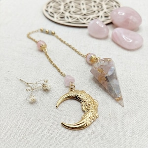 Peut inclure: Un pendule composé d'une pierre pointue translucide avec des éclats dorés, fixé à une chaîne dorée. La chaîne comprend de petites perles roses et une breloque en forme de croissant de lune doré avec un motif floral en relief sur sa surface.