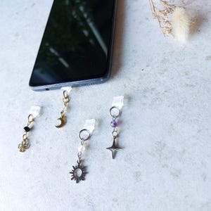 Könnte beinhalten: Vier Handy-Charm-Accessoires mit dekorativen Anhängern. Ein Anhänger zeigt ein goldenes Kleeblatt, ein anderer einen goldenen Mond, ein dritter eine Sonne und der letzte einen Stern. Jeder Anhänger ist an einem transparenten Stecker befestigt.
