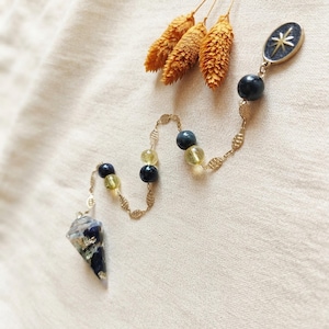 May include: A pendulum featuring a conical resin weight with dark blue and gold-toned inclusions, suspended from a gold-toned chain. The chain is accented with alternating black and translucent yellow beads, ending in an oval charm with a star design.