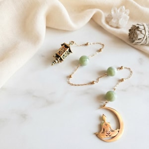 Peut inclure: Un pendule en métal doré composé d'une petite breloque lanterne, de trois perles lisses vert pâle et d'un pendentif croissant de lune avec la silhouette découpée d'une personne en position de méditation assise avec un symbole Om sur la poitrine.