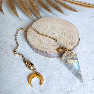 Op de afbeelding: Een goudkleurige pendel met een helder, puntig kristal en gouden bladaccenten. De pendel hangt aan een gouden ketting en heeft een halve maan bedel en een gouden bol. Het kristal ligt op een houten schijf.