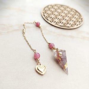 May include: A gold-toned chain pendulum featuring two pink spherical beads, a small gold heart charm, and a conical resin pendant containing pink material and gold leaf flakes, suspended from a delicate metal link chain.