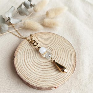 Peut inclure: Un collier à chaîne dorée présentant une disposition verticale de pendentifs. Le design comprend un petit maillon doré, une perle sphérique blanche lisse, une perle translucide à facettes et une breloque en forme de goutte dorée polie.