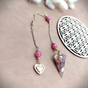 Könnte beinhalten: Ein goldfarbenes Pendel mit einem klaren, spitz zulaufenden Kristall und Goldblatt-Akzenten, verbunden durch eine Kette mit rosa Edelsteinperlen und einem herzförmigen Anhänger. Ein dekoratives Holzstück befindet sich im Hintergrund.