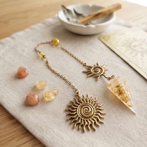 Peut inclure: Un pendule à chaîne en métal doré avec une breloque en forme de soleil au centre en spirale et un poids en résine pointu et transparent contenant des flocons d'or. L'ensemble comprend deux petites pierres lisses orangées et deux pierres jaunes.