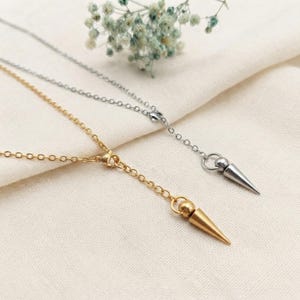 Puede incluir: Dos collares estilo lariat, uno en tono dorado y otro en tono plateado, cada uno con una cadena fina y un colgante cónico liso suspendido de un pequeño anillo circular. Los collares están dispuestos para mostrar los dijes metálicos puntiagudos.